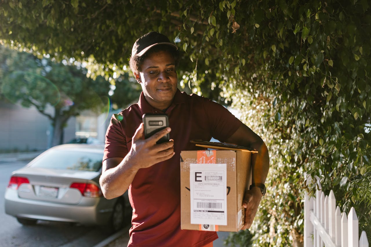 A male courier delivering a package in a sunny urban neighborhood while using a smartphone.